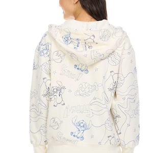 May include: Cream-colored hoodie featuring a blue line art design of Stitch, planets, and stars. The word "Stitch!" is repeated throughout the design. The hoodie has a drawstring hood and ribbed cuffs and hem.