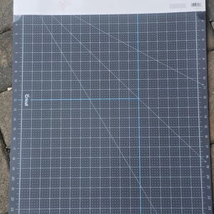 May include: A gray Cricut cutting mat with a grid pattern and measurements in inches. The mat has a white border at the top with the Cricut logo and product information. The grid lines are white and blue, and the mat is designed for crafting.