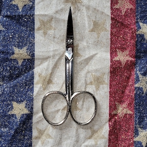 May include: Silver cuticle scissors with a pointed tip and two rounded loops. The scissors are lying on a white fabric with a red and blue star pattern.