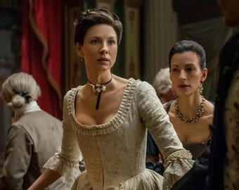 May include: Two women in period dress, one wearing a white gown with a black choker necklace, the other wearing a dark dress with a gold necklace. The woman in the white gown is looking to the right, while the woman in the dark dress is looking at the camera.