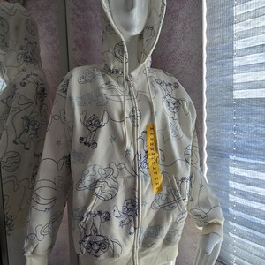 May include: Cream-colored zip-up hoodie featuring a blue cartoon character design. The hoodie has a drawstring hood, front pockets, and a yellow tag. The design includes the character, planets, and stars.