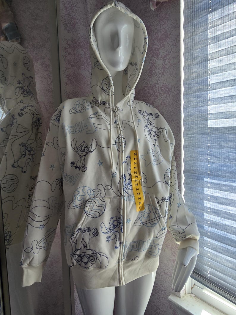 May include: Cream-colored zip-up hoodie featuring a blue cartoon character design. The hoodie has a drawstring hood, front pockets, and a yellow tag. The design includes the character, planets, and stars.