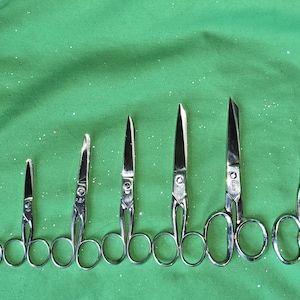May include: Seven silver scissors of varying sizes, arranged in a row on a green fabric background.