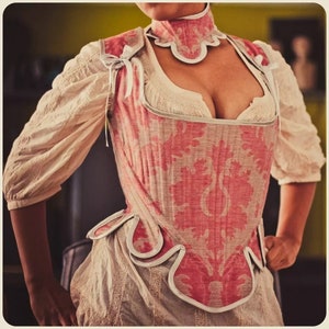 Op de afbeelding: Een roze en witte corset met een bloemenpatroon. Het corset heeft een gegolfde halslijn en een witte kantversiering.