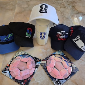May include: Four baseball caps and a white water bottle featuring FIFA World Cup 26 branding, alongside two square fabric mats with a central pink soccer ball graphic. The caps display various logos including Philadelphia, USA, and the FIFA 26 emblem.