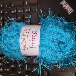 Pode incluir: Um novelo de fio azul turquesa com textura felpuda. O fio é etiquetado como "Moda Dea Prima."