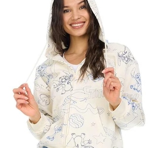 May include: Cream-colored zip-up hoodie with a hood and blue line art illustrations. The design features cartoon characters, planets, and stars. The word "Stitch" is also visible on the front of the hoodie.