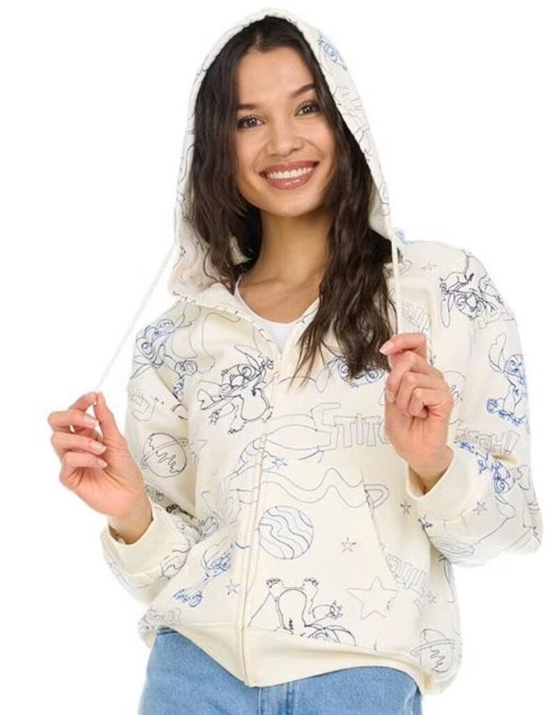 May include: Cream-colored zip-up hoodie with a hood and blue line art illustrations. The design features cartoon characters, planets, and stars. The word "Stitch" is also visible on the front of the hoodie.