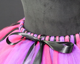 May include: A close-up of a colorful tutu skirt. The skirt features layers of pink and purple tulle, accented with black stripes. A black satin ribbon is tied in a bow at the waist. The tutu is displayed on a black mannequin.