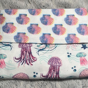 May include: A folded white pillowcase with a seashell pattern in pink, blue, and yellow on the top half. The bottom half features a pattern of purple jellyfish, teal waves, and pink sea stars.