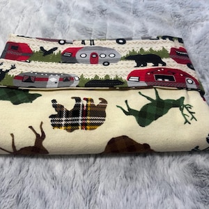 May include: Folded fabric with a camping theme. The top layer features red and gray camper illustrations, black bears, and deer. The bottom layer has deer and bear silhouettes, and plaid patterns in green, brown, and cream.
