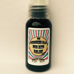Könnte beinhalten: Eine kleine, dunkelbraune Flasche mit einer durchsichtigen Plastik-Pumpenaufsatz. Die Flasche hat ein weißes Etikett mit roten, grünen und weißen Streifen und dem Text "The Medicine Man Bug Bite Relief" in Schwarz.