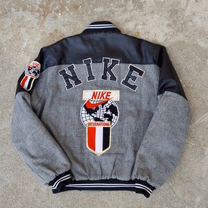 May include: A gray and black varsity jacket with a large "NIKE" embroidered on the back. The jacket also features a smaller "NIKE INTERNATIONAL" logo with a globe and swoosh.