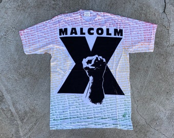 Pode incluir: Camiseta branca com um "X" preto e um punho levantado. O "X" é impresso com as palavras "MALCOLM" e "By Any Means Necessary" repetidas em vermelho e amarelo.