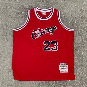 May include: Red basketball jersey with white trim and black lettering. The jersey says "Chicago" and "23" on the front. The jersey has a patch that says "Hardwood Classics 1983-84".