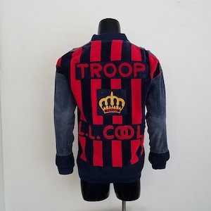 May include: A navy blue and red striped jacket with a crown and "TROOP" embroidered on the back. The sleeves are denim.