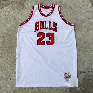 May include: White basketball jersey with red trim and the words "BULLS" and "23" in red. The jersey has a tag that says "1984-85" and "SWINGMAN"