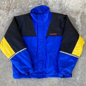 May include: A blue, black, and yellow Polo Sport jacket. The jacket has a high collar, a front zipper, and the words "POLO SPORT" in orange. The sleeves are black with yellow accents and reflective stripes.