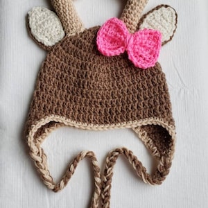 May include: A brown crocheted deer hat with white ears and antlers. The hat has a pink bow on top and a brown and white braided tie.