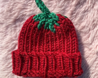 May include: A handmade red knit hat with a green stem and leaves, resembling a strawberry. The hat is ribbed and has a folded brim. The hat is made of a soft, textured yarn.