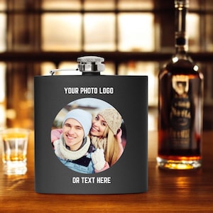 Pode incluir: Frasco de metal preto com um inserto de foto redonda de um casal com roupas de inverno. O texto "YOUR PHOTO LOGO" e "OR TEXT HERE" está impresso no frasco.