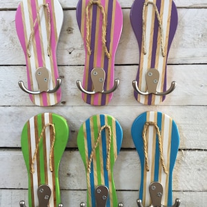 May include: Six wooden flip-flop shaped hooks with double hooks. The flip-flops are painted in various colors with white stripes. The hooks are mounted on a white wooden wall.