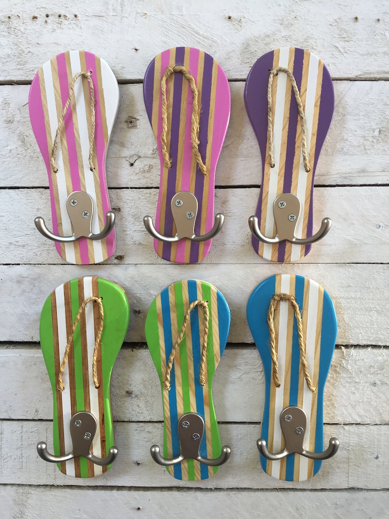 May include: Six wooden flip-flop shaped hooks with double hooks. The flip-flops are painted in various colors with white stripes. The hooks are mounted on a white wooden wall.