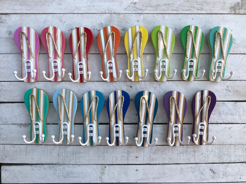 May include: A set of 16 colorful flip-flop shaped hooks with white rope detailing and three hooks on each. The hooks are arranged in two rows of eight on a white wood background.