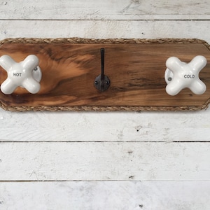 May include: A wooden towel rack with a rope border and two white porcelain knobs labeled "HOT" and "COLD". A single black metal hook is centered on the rack.