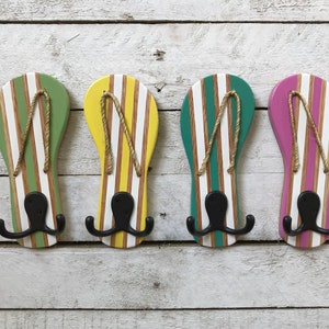 May include: Four wooden flip-flop shaped hooks painted in green, yellow, teal, and pink with white stripes. Each hook has a black double hook.