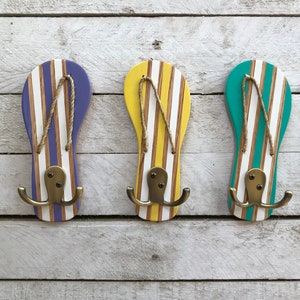 May include: Three wooden flip-flop shaped hooks painted in purple, yellow, and green with white stripes. Each hook has two brass hooks.