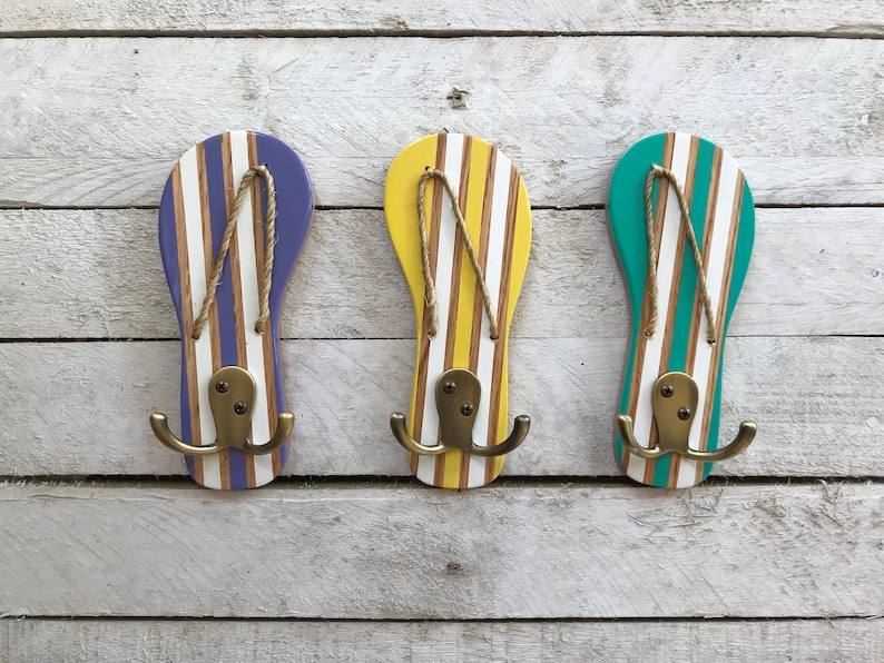 May include: Three wooden flip-flop shaped hooks painted in purple, yellow, and green with white stripes. Each hook has two brass hooks.
