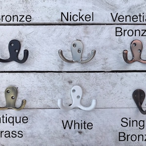 May include: Six different double hooks in various finishes, including bronze, nickel, Venetian bronze, antique brass, white, and single bronze. The hooks are displayed on a white wooden background.