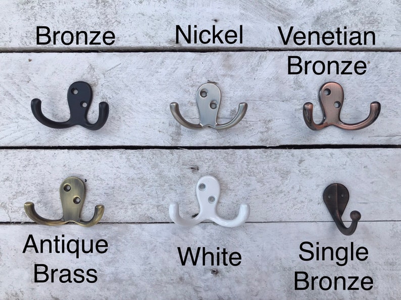 May include: Six different double hooks in various finishes, including bronze, nickel, Venetian bronze, antique brass, white, and single bronze. The hooks are displayed on a white wooden background.