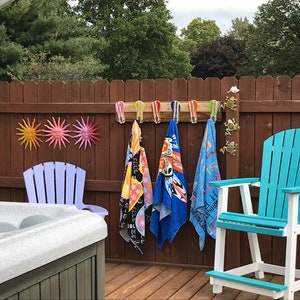 May include: A wooden rack with hooks holds four colorful beach towels. The rack is mounted on a brown wooden fence. There are three metal sun decorations on the fence. A blue and white Adirondack chair is in the foreground. A hot tub is in the background.