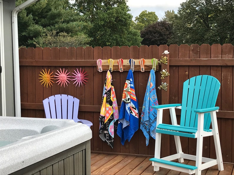 May include: A wooden rack with hooks holds four colorful beach towels. The rack is mounted on a brown wooden fence. There are three metal sun decorations on the fence. A blue and white Adirondack chair is in the foreground. A hot tub is in the background.