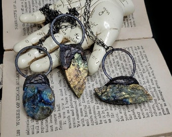 Puede incluir: Tres colgantes artesanales de piedras preciosas con detalles en metal oscuro. Cada colgante presenta una piedra iridiscente única en tonos azules, dorados y verdes, suspendida de una cadena. Los colgantes descansan sobre un libro abierto.