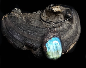Puede incluir: Collar con colgante artesanal con una gran piedra de labradorita ovalada con tonos azules y verdes. La piedra está engastada en un marco de cobre rústico y texturizado y cuelga de un cordón negro. El colgante descansa sobre una superficie de madera oscura y texturizada.