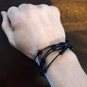 May include: A black bracelet featuring a design that mimics barbed wire. The accessory consists of multiple thin, dark, flexible strands wrapped around the wrist, with small, knotted, spike-like protrusions spaced evenly along the length of the cord.