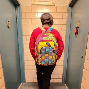 May include: A person wearing a red long-sleeved shirt and black pants is carrying a multi-colored backpack with a yellow, blue, and purple design. The backpack has a zippered pocket on the front.