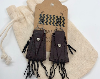 May include: A pair of brown leather earrings with fringe details. The earrings are hanging from a brown paper tag with the text "Handmade" and "Rescue" on it. The earrings are packaged in a small white drawstring bag.