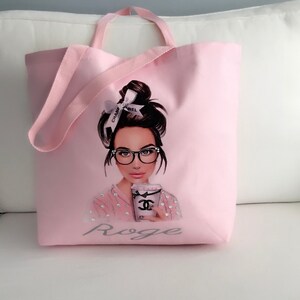 May include: Pink canvas tote bag with a graphic of a woman wearing glasses and holding a coffee cup with a Chanel logo. The bag has a gray text that says "Roge".