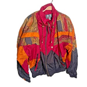 Puede incluir: Chaqueta cortavientos vintage y colorida con una mezcla de paneles rojos, naranjas, marrones y negros. La chaqueta tiene cremallera frontal, cuello alto y capucha con cordón. La chaqueta está colgada de una percha de madera.
