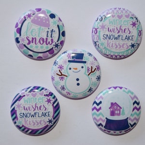 May include: Five circular pinback buttons featuring winter themes. One displays a snowman, another a snow globe with a house, and two feature the text "winter wishes SNOWFLAKE kisses". The final button shows a hat and the text "let it snow".