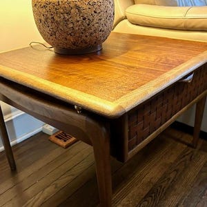 May include: A mid-century modern side table with a rectangular top and woven drawer. The table is made of wood with a warm, natural finish. A lamp with a cork-like base sits on top of the table.