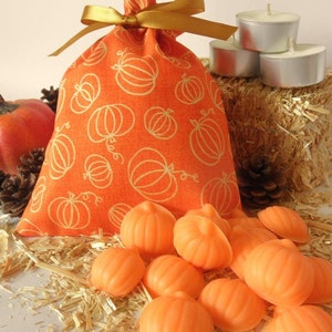 May include: Orange fabric bag with a gold ribbon and a pumpkin pattern. The bag is filled with small orange pumpkin-shaped candles. The candles are sitting on a bed of straw.