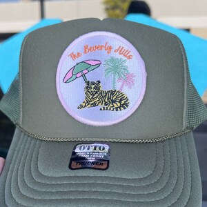 May include: Olive green trucker hat with a circular patch featuring a tiger, palm trees, and the text "The Beverly Hills." The hat has a foam front and mesh back. The hat also has a sticker that says "World Famous Foam Front".