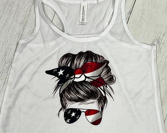 Pode incluir: Top branco com design de costas de nadador. A frente apresenta um gráfico do rosto de uma mulher com um coque bagunçado, óculos de sol e uma bandana, todos decorados com a bandeira americana. O top é feito de um material macio e leve.