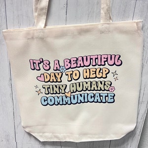 Puede incluir: Una bolsa de lona blanca con el texto "It's a beautiful day to help tiny humans communicate" en una fuente retro con acentos coloridos y pequeños gráficos de flores y corazones.