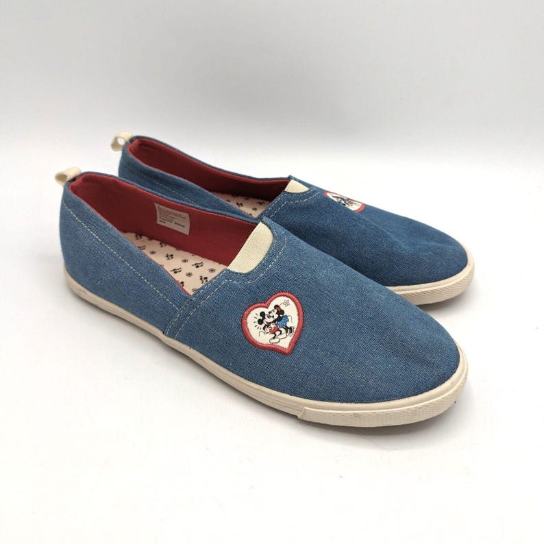 May include: Denim slip-on shoes with a white sole and red lining. Each shoe has a heart-shaped patch with an embroidered Mickey Mouse design. These casual shoes are suitable for everyday wear.